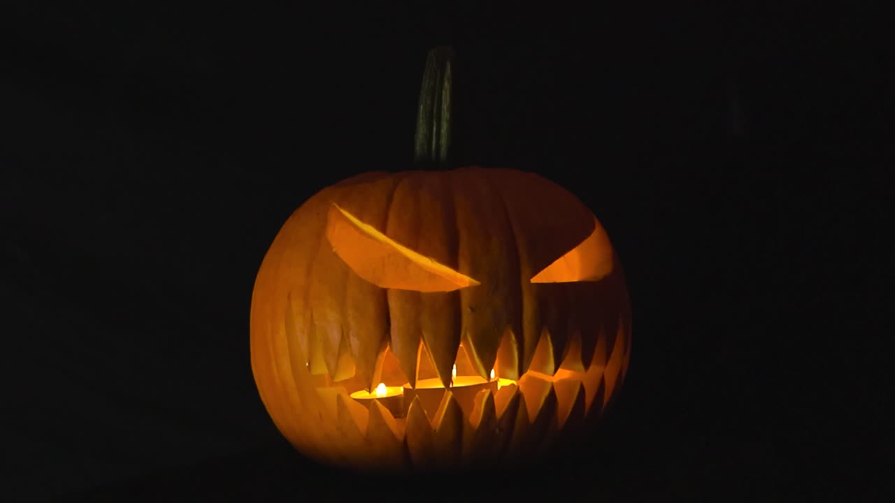 Scary carved orange colored Halloween jack o lantern pumpkin with candles burning inside that is placed in front of a black studio background, smoke visible in the foreground