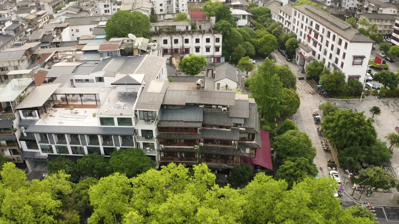 Una processione dall'alto si addentra tra le chiome color smeraldo in un intricato quartiere sul fiume, con edifici piastrellati e vicoli verdeggianti che si aprono sotto cieli velati da rocce carsiche, nella serenità senza tempo di Yangshuo.
