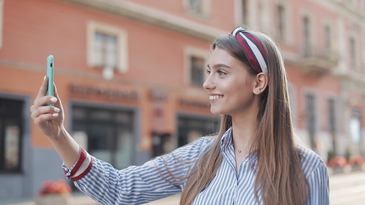 Giovane donna sorridente che saluta con la mano mentre si scatta un selfie in una strada di città.