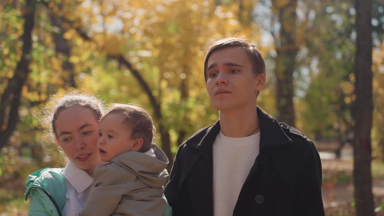 Famiglia caucasica in un parco autunnale che abbraccia un bambino piccolo, la madre coccola il figlio mentre il padre osserva, tra foglie dorate e calda luce solare, con sorrisi gentili, un intimo momento coniugale di legame e genitorialità.
