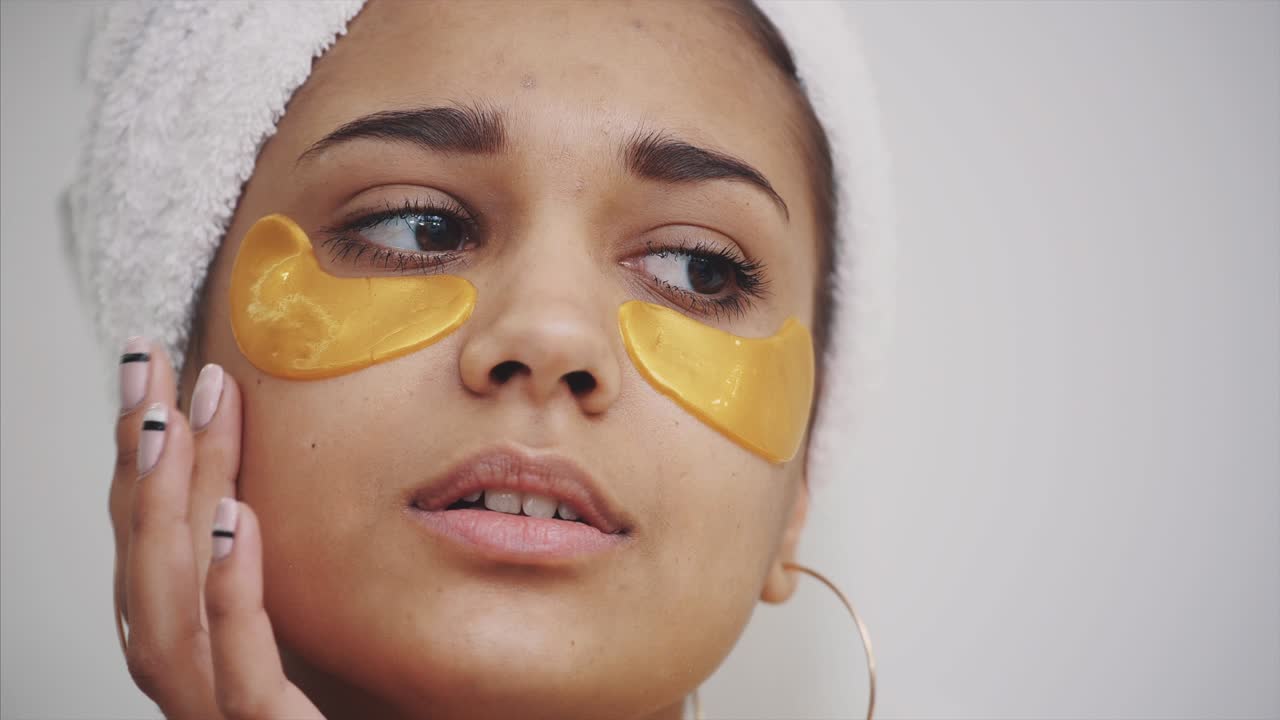 Closeup portrait of beautiful woman after bath with towel on head puts patches under the eyes from wrinkles and dark circles. Attractive female in bathrobe makes beauty treatments on the face at home.