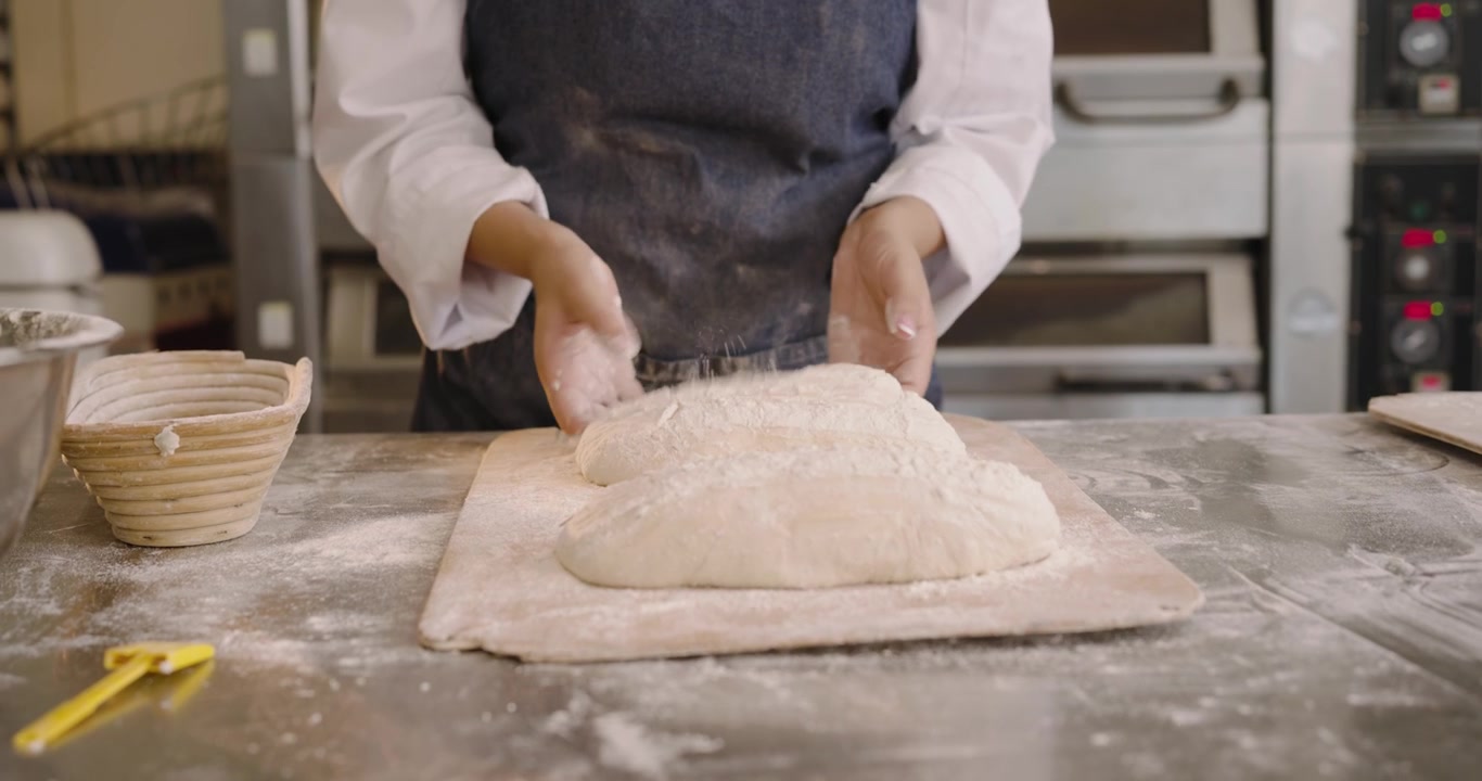 Panettiere di mezza età con grembiule al bancone, solleva un cesto mostrando l'impasto che modella pagnotte sulla pala per la cottura.