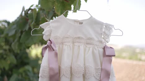 elegant baby dress with lace details and pink ribbons, hanging on a hanger against a natural background, perfect for a christening or special occasion