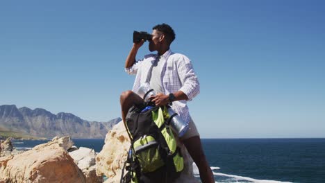 Afroamerikanischer-Mann,-Der-Mit-Ferngläsern-Wandert-Und-Auf-Einem-Felsen-An-Der-Küste-Sitzt