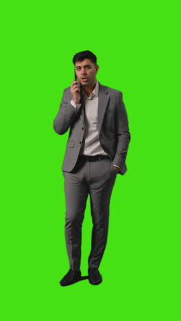 Vertical-Video-Of-Businessman-Talking-On-Mobile-Phone-Walking-Into-Frame-Against-Green-Screen-Background