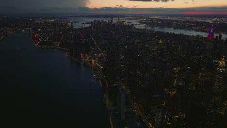 Fly-above-East-River-bank-in-evening.-Tilt-up-reveal-cityscape-against-colourful-twilight-sky.-Manhattan,-New-York-City,-USA