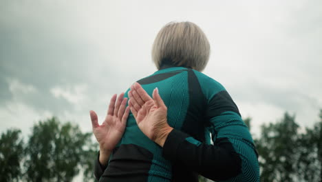 vista posterior de cerca de una mujer de mediana edad sentada en la postura de sukhasana, practicando yoga al aire libre, el fondo muestra árboles borrosos alineados bajo un cielo nublado