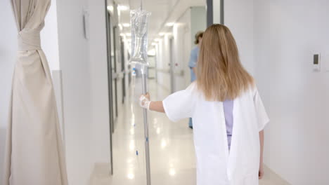 rear view of caucasian girl patient walking with drip stand in hospital, copy space, slow motion