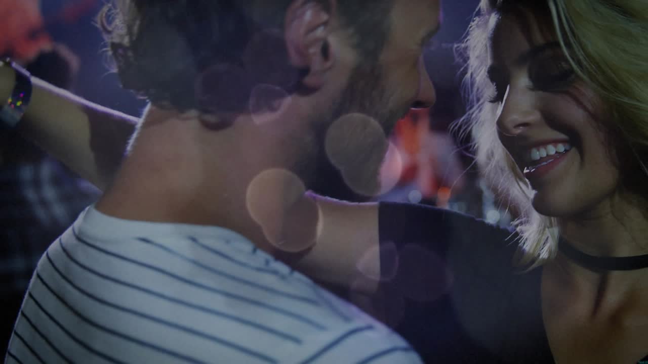 Couple sharing smile, creating light flares in club woman looping arm, drawing man closer playfully