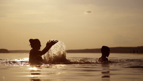 woman splashing water over the boy on the evening river background. silhouette of a mother and her son spending time together in water at sunset.