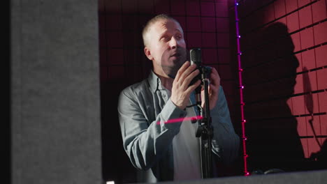 a man in a blue long-sleeve shirt and white inner shirt passionately sings into a microphone within a red soundproof studio, the lighting casting dramatic shadows and enhancing the atmosphere