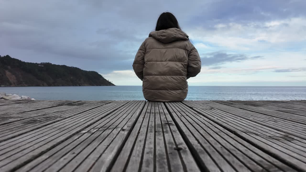 Slow-motion back view of a woman seated alone facing the sea on a wooden deck