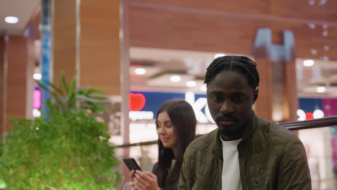 Angry man turns head to glance at woman smiling while holding phone in blurred mall setting with bright lights, green indoor plants, and wooden walls