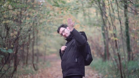 un hombre europeo barbudo con gafas afiladas, de pie en un bosque mixto de otoño, apuntando y llamando con entusiasmo a los espectadores más cerca, invitándolos a unirse a un viaje de exploración de la naturaleza