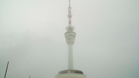 huge tall radio tower with a long antenna red and white coming out of a building in mountains high point above inside the clouds and a mysterious mist it is giving thriller horror vibes cinematic sky