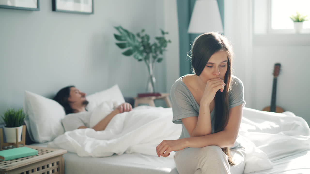 Sad Woman Sitting in Bed Next to Sleeping Man