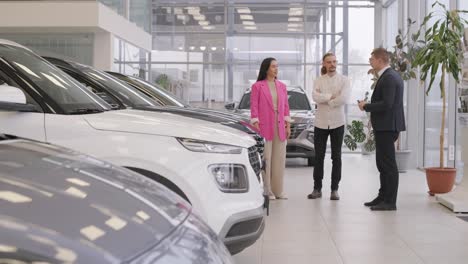 young car salesman showing to young couple new automobile at dealership salon.