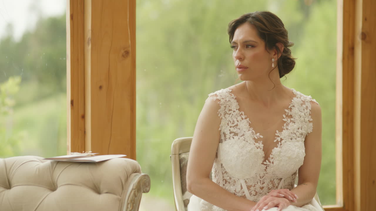 A bride sits on a sofa, calmly gazing out the window overlooking the countryside. The wedding invitation rests beside her, in an intimate, elegant, and thoughtful scene.