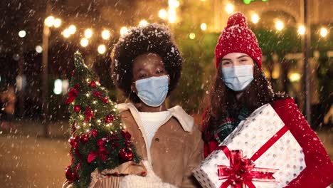 felices amigas caucásicas y afroamericanas sosteniendo regalos y sonriendo a la cámara en la calle mientras nieva en navidad