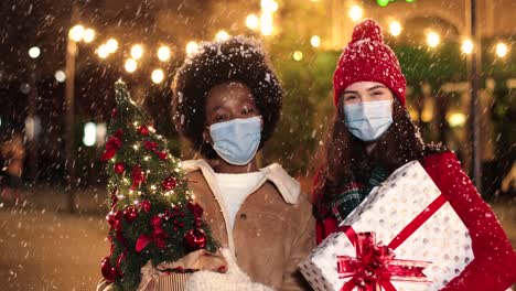 glückliche kaukasische und afroamerikanische freundinnen, die geschenke halten und auf der straße in die kamera lächeln, während es in weihnachten schneit