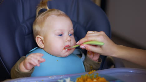 blue-eyed baby having meals. kid takes some food with his hand and mom wants to clean it.