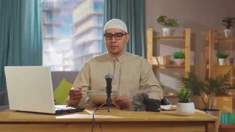 muslim man in a skullcap and wearing glasses records a podcast while sitting in front of a microphone in living room