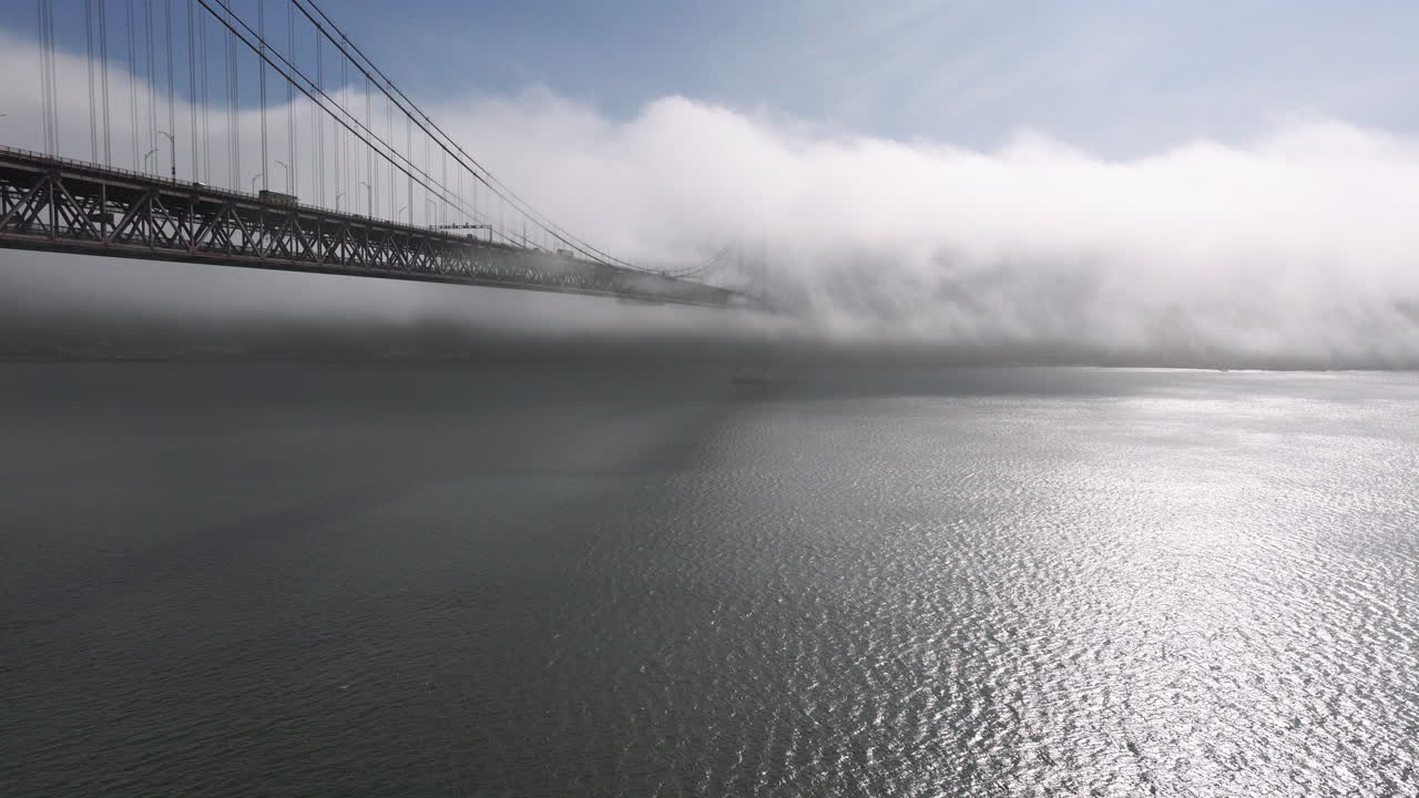 Aerial drone shot towards the 25th April Bridge in Lisbon, Portugal, Europe. Sunny and bright with clouds, fog, mist, covering the bridge. Shot in ProRes 422 HQ