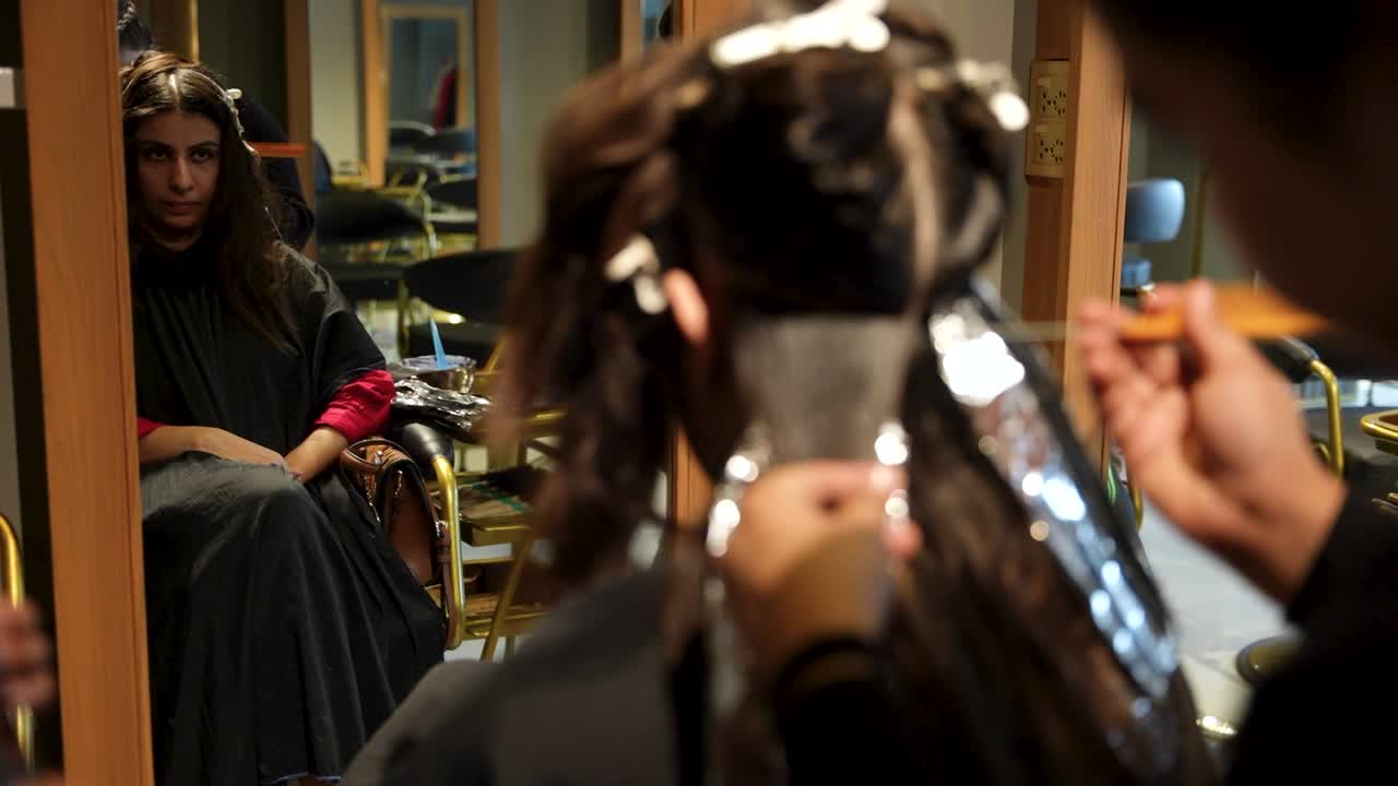 Donna seduta su una poltrona da parrucchiere mentre l'hairdresser applica il colore su ciocche di capelli avvolte in stagnola riflesse nello specchio