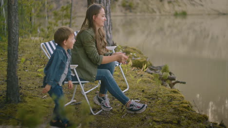 Una-Mujer-Con-Un-Niño-Pequeño-Descansa-En-La-Costa-De-Un-Pintoresco-Lago-En-El-Bosque-Arrojando-Piedras-Al-Agua,-Diversión-Familiar-Feliz-En-La-Naturaleza