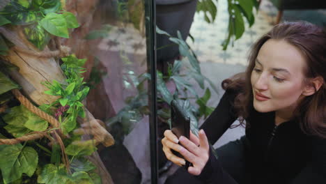 smiling woman with blond hair crouches to photograph lush indoor greenery behind glass wall, her reflection faintly visible on surface, creating soft layered composition with plants