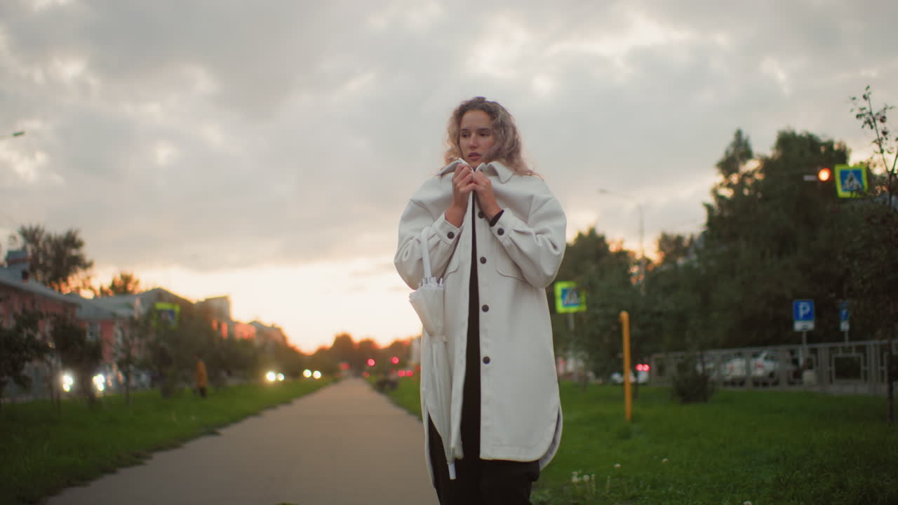 Cold lady covering herself with white coat while small dog passes by on green parkway during chilly evening with blurred background featuring moving cars, urban streetlights, buildings and trees
