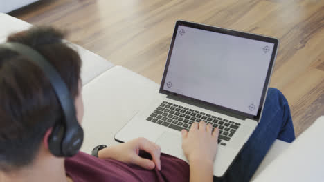 Asian-male-teenager-wearing-headphones-and-using-laptop-in-living-room