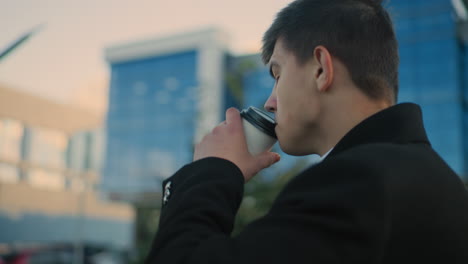 man in black coat focusing on phone while sipping tea in an outdoor urban setting featuring blurred greenery and modern glass building in background