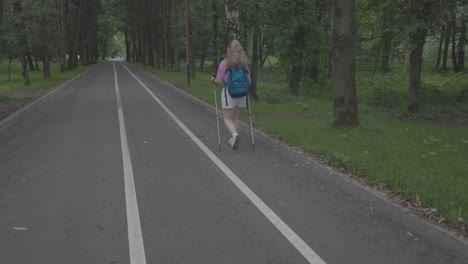 A-shot-from-the-back,-a-girl-walking-on-an-asphalt-road-in-a-forest,-using-walking-sticks,-surrounded-by-trees-and-green-herbs,-summer