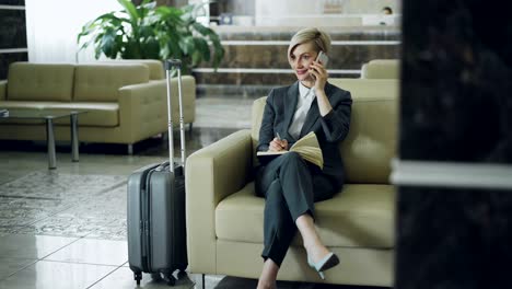 attractive businesswoman sitting in armchair in hotel lobby talking on mobile phone and writing in notepad smiling and looking aside. business, travel and people concept