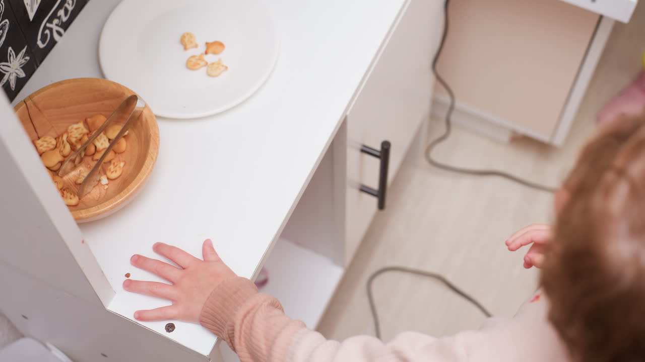 kid in pink sweater carefully using fork to transfer fish-shaped snacks from wooden bowl onto plate, standing at white counter in bright kitchen setting then left with the plate