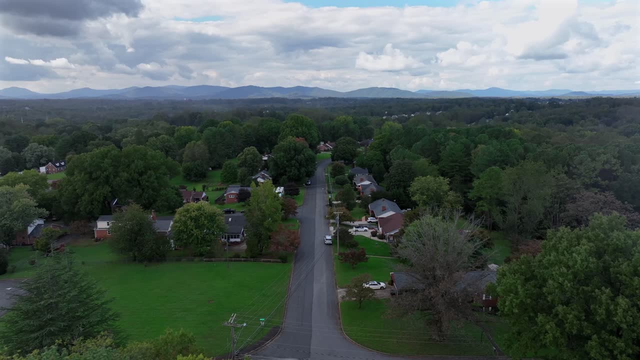 American suburb neighborhood with straight street during cloudy day in autumn. Housing. Area with parking cars along green tree road. Mountain silhouette at horizon. Aerial landing shot.