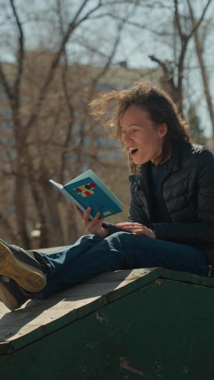 Close up of woman reading book outdoors on wooden platform with wind blowing hair. Book has colorful design subject wears black jacket and blue jeans. Sunny day background includes trees