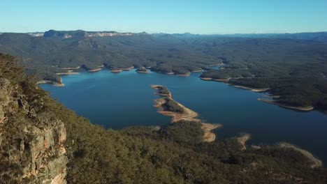 aerial: drone shot slowly tracking alongside a rocky mountain towards a large blue lake surrounded by australian bush land