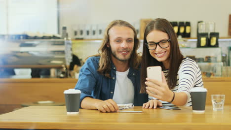 caucasian man and woman friends making a video call using smartphone sitting at a table in a cafe