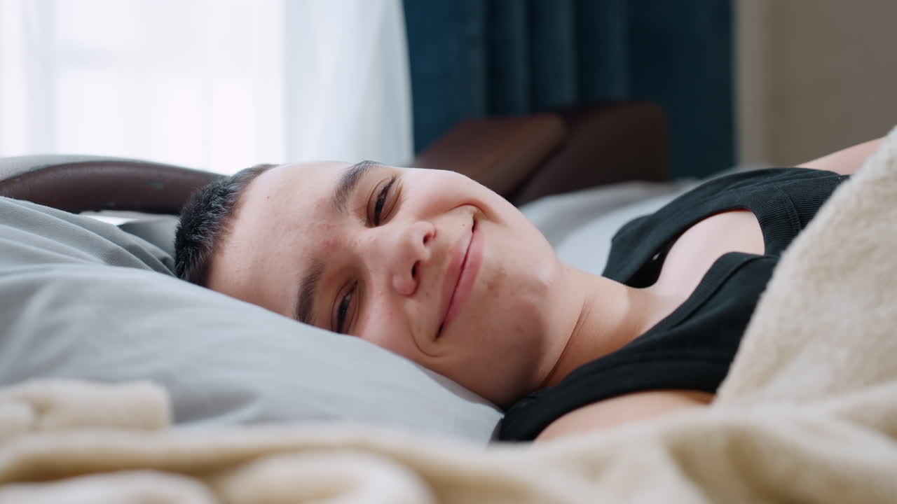 Close up kid resting on pillow under beige blanket wearing black singlet gently opens eyes and shows warm smile creating cozy serene atmosphere of comfort