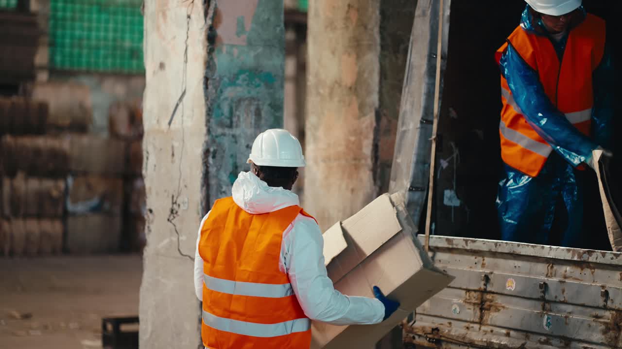 A man with Black skin in a white protective uniform in an orange vest accepts waste paper given to him by a brunette girl from a truck and stores it at the Large Waste Recycling and Sorting Plant