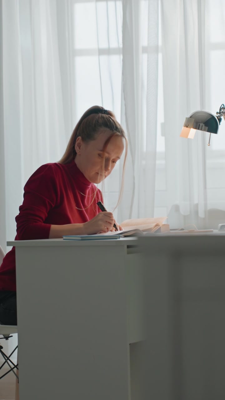Woman in red top writes or draws on open book at white desk. Scene features natural light from sheer curtains. Close up view shows consistent engagement with study materials