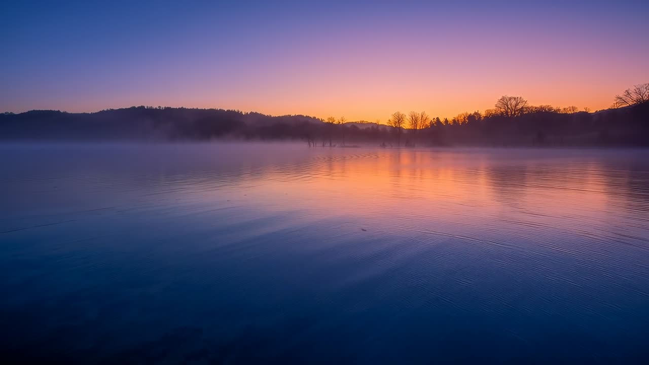 Reflecting dawn colors, forming ripples as sun rising, calm lake surface with mist and tree line