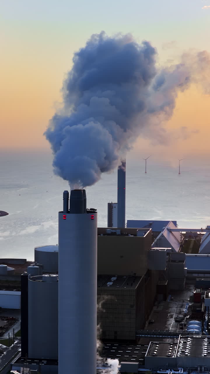 Aerial drone view of Amager Bakke power plant chimney releasing smoke, with offshore wind turbines standing in the sea behind it in Copenhagen, Denmark. Vertical