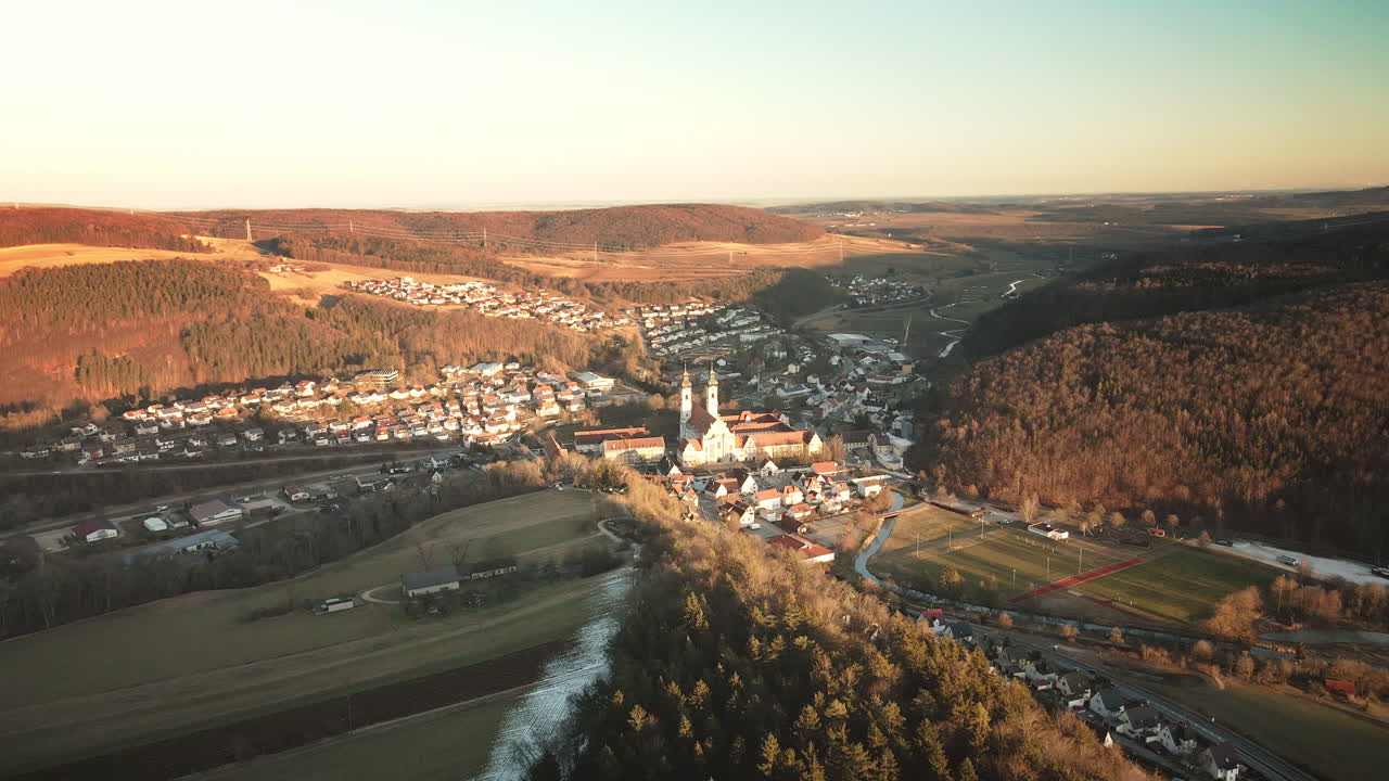 Drone advances forward over Zwiefalten's quaint rooftops toward hilltop monastery towers amid autumn hills, fields and river in warm Swabian Alb sunset. Timeless baroque panorama