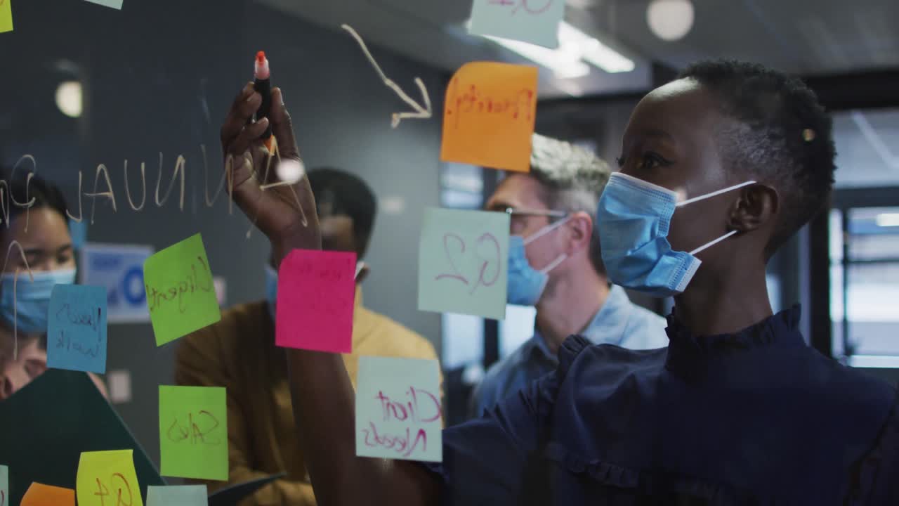 African american woman wearing face mask writing with marker pen on on glass board at modern office