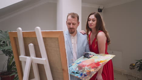 woman leans on artist shoulder in studio. languid socialite in red gown hugs art master drawing lady painting in art workshop. muse inspires painter