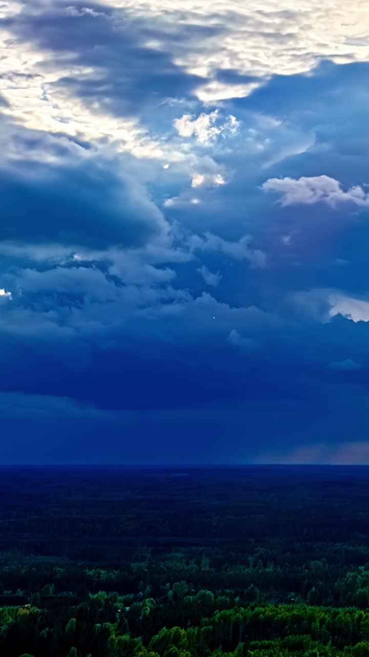 Vertical timelapse of dramatic storm clouds rolling over dense green forest in Latvia at sunset, filling deep blue sky with majestic cumulonimbus formations