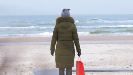 young woman walking along the beach in winter overcast day, red bag in right hand, handheld medium shot